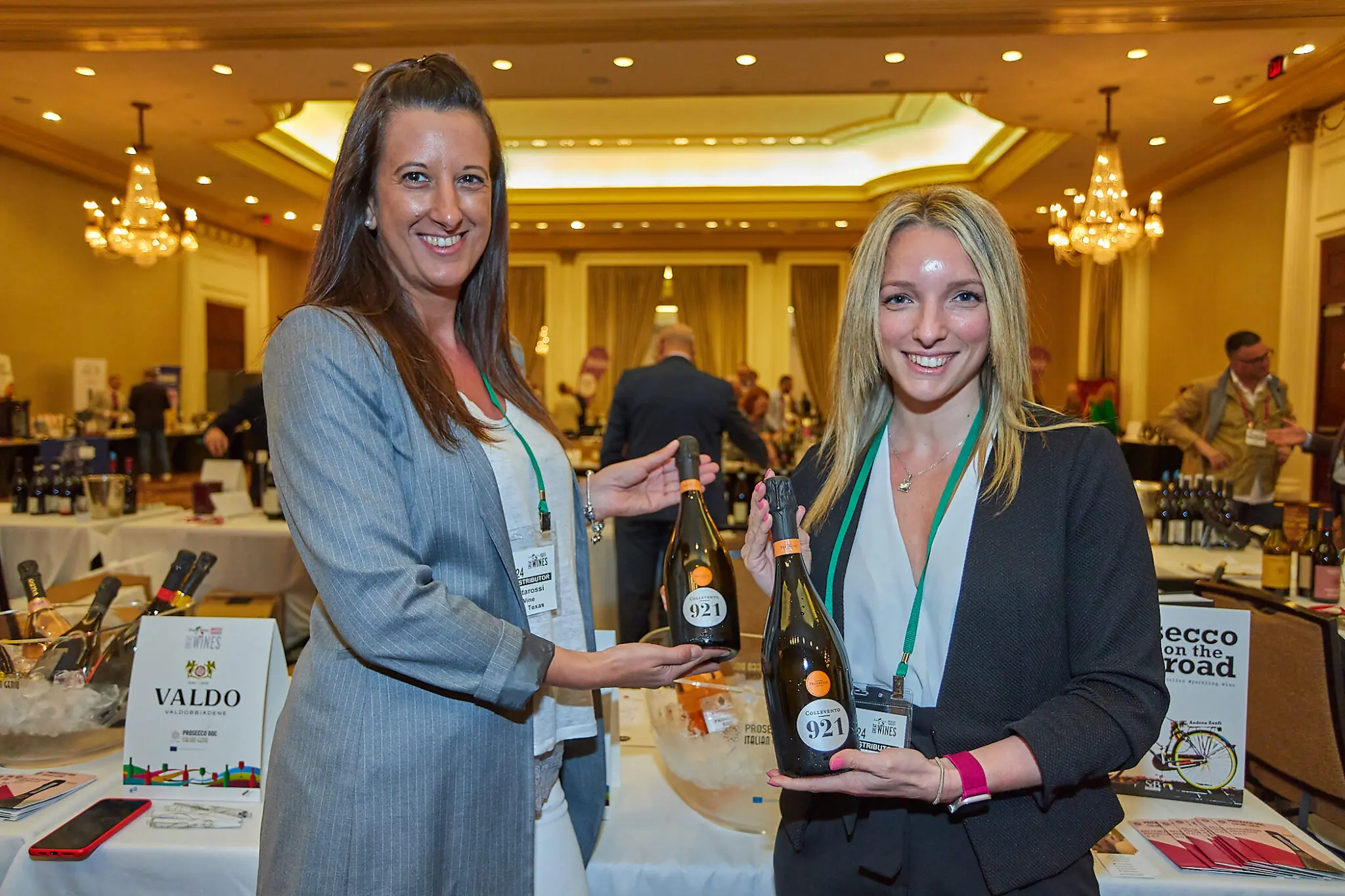 Two women smiling and holding wine bottles at an indoor event. Tables with more bottles and people are visible in the background.