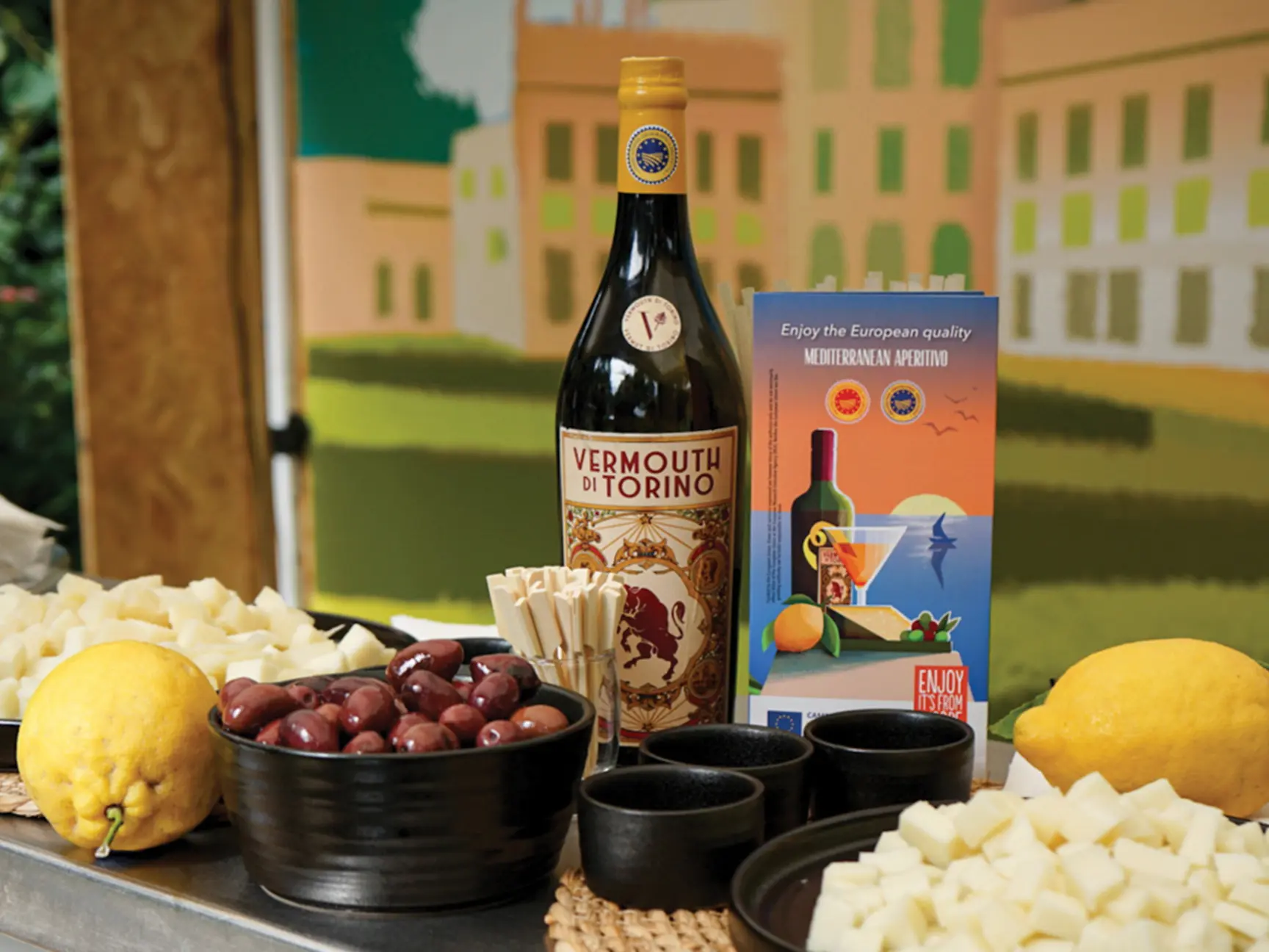 A bottle of Vermouth di Torino and its box sit on a table with bowls of olives, cheese cubes, and lemons, in front of a colorful illustrated backdrop.